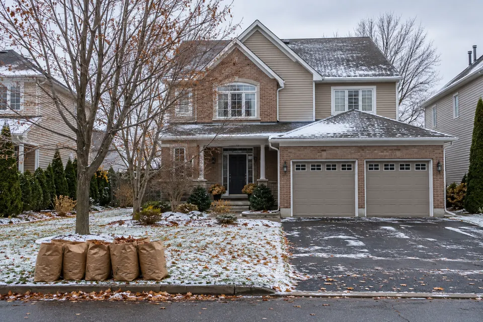 A two-storey brick and siding house in an Ottawa suburb in November, clean white eavestroughs and paper yard-waste bags of leaves at the curb
